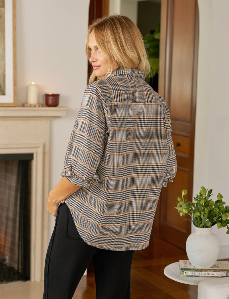 A woman with blonde hair stands indoors, looking over her shoulder. She wears the Frank & Eileen Eileen Button Up Shirt F25 in a relaxed, casual California style with black pants. Behind her are a white fireplace, candles, wooden doors, and a plant on a round table.