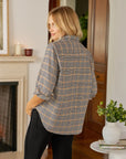 A woman with blonde hair stands indoors, looking over her shoulder. She wears the Frank & Eileen Eileen Button Up Shirt F25 in a relaxed, casual California style with black pants. Behind her are a white fireplace, candles, wooden doors, and a plant on a round table.