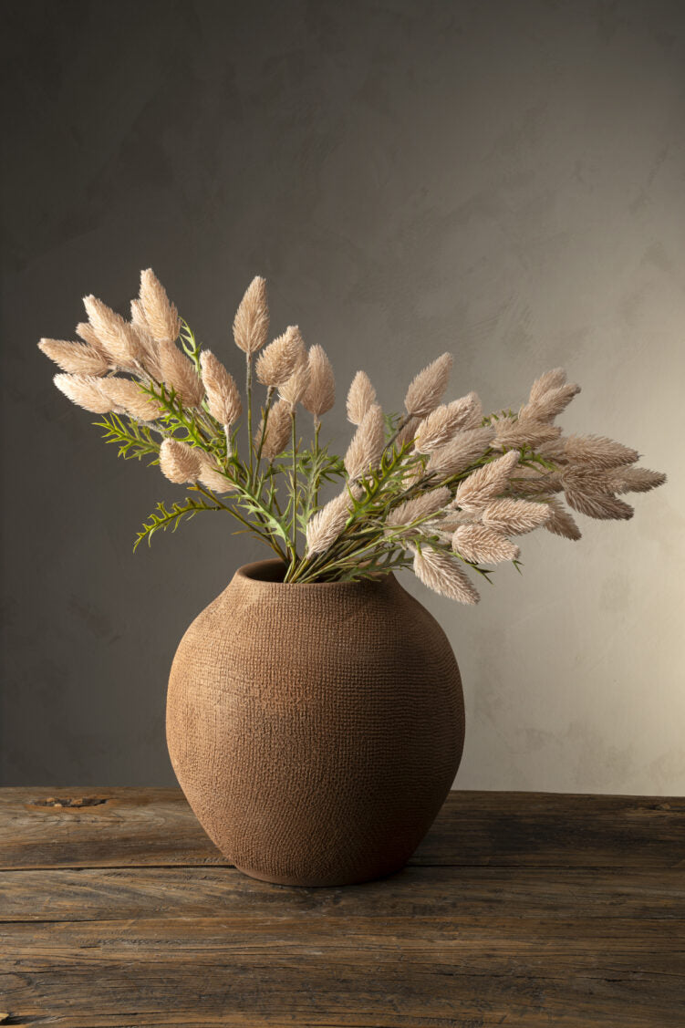 The Indaba Pussytail Grass Spray, featuring fluffy beige dried stems and green faux plants, is arranged in a round textured brown vase on a rustic wooden table set against a softly lit neutral background.