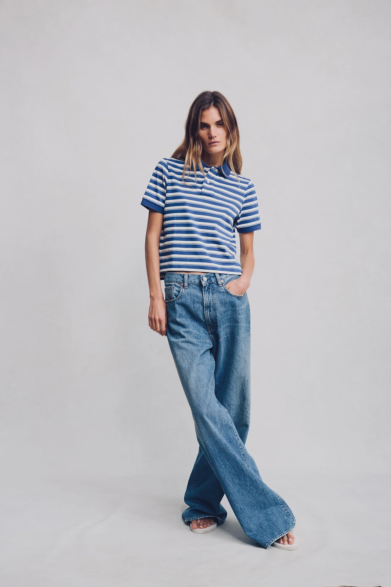A woman stands against a plain background wearing Denimist's SHRUNKEN POLO, a blue and white striped cotton t-shirt, paired with loose-fitting blue jeans. Her left hand rests in her pocket while her right arm is relaxed at her side.