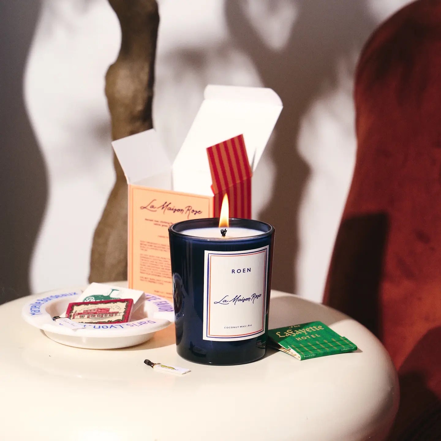 A Roen Candle by Faire, in a dark blue jar made from a coconut soy wax blend, sits on a round table. An open box is behind it, with matches on a plate and a green tea bag nearby. A brown textured sculpture complements the serene ambiance of Casa Polanco.