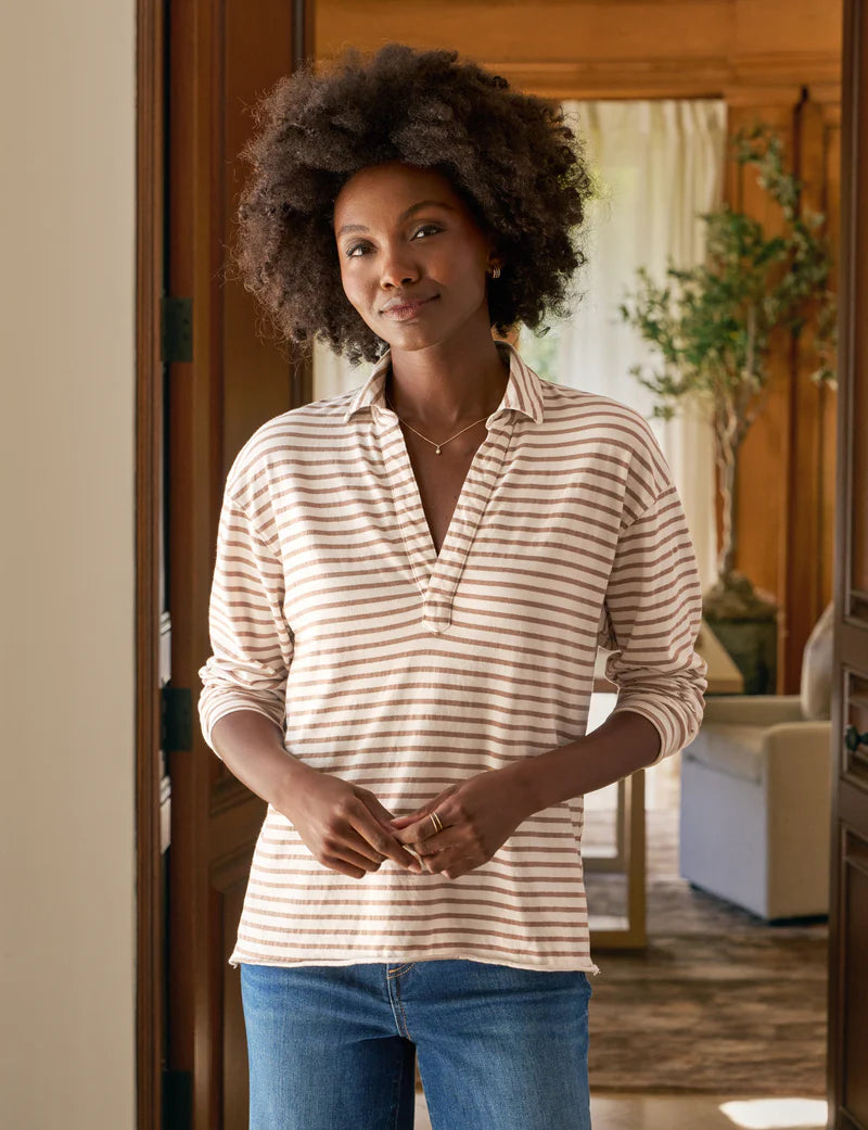A woman with curly hair wears the Frank & Eileen Patrick Popover Henley, a beige and white striped cropped long-sleeve shirt, paired with blue jeans. She stands indoors, smiling gently, with a blurred living room in the background.