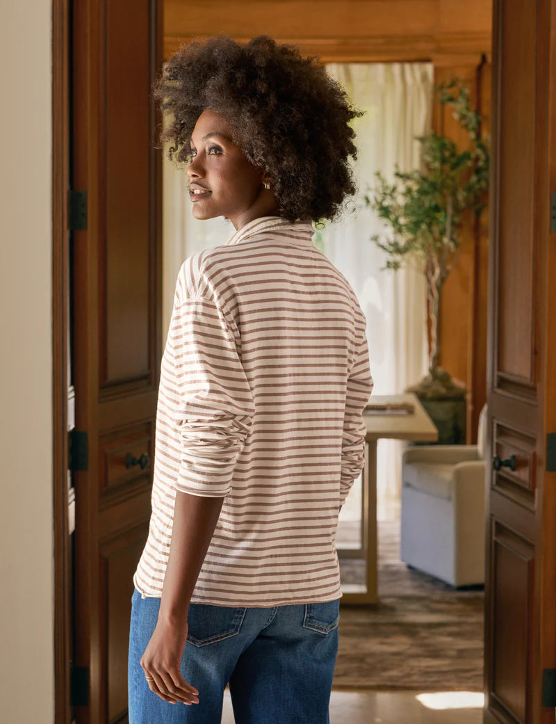 A woman with curly hair stands indoors, sunlight streaming behind her, wearing the Patrick Popover Henley by Frank & Eileen—a striped, cropped long-sleeve shirt—paired with jeans and looking over her shoulder.