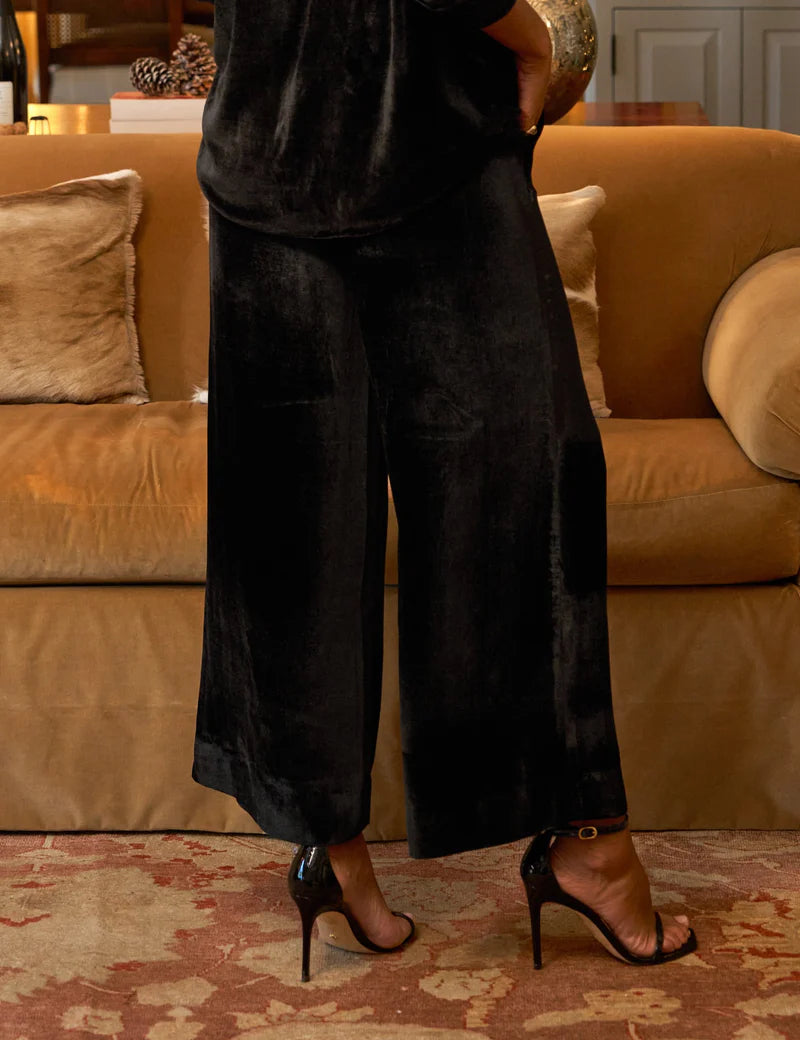 A person wears the Frank & Eileen Tuscany Getaway Maisie Pant, featuring a loose black velvet wide-leg silhouette, paired with black heels while standing before a tan sofa with plush cushions and beige arms on a patterned rug.