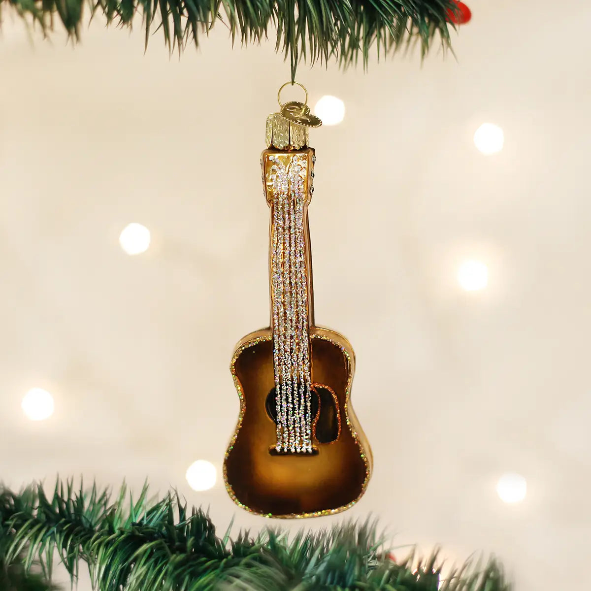 The Old World Ornaments guitar-shaped ornament by Faire, featuring brown and gold details with sparkling strings, hangs on a green Christmas tree branch among other decorations against a backdrop of warm white lights.