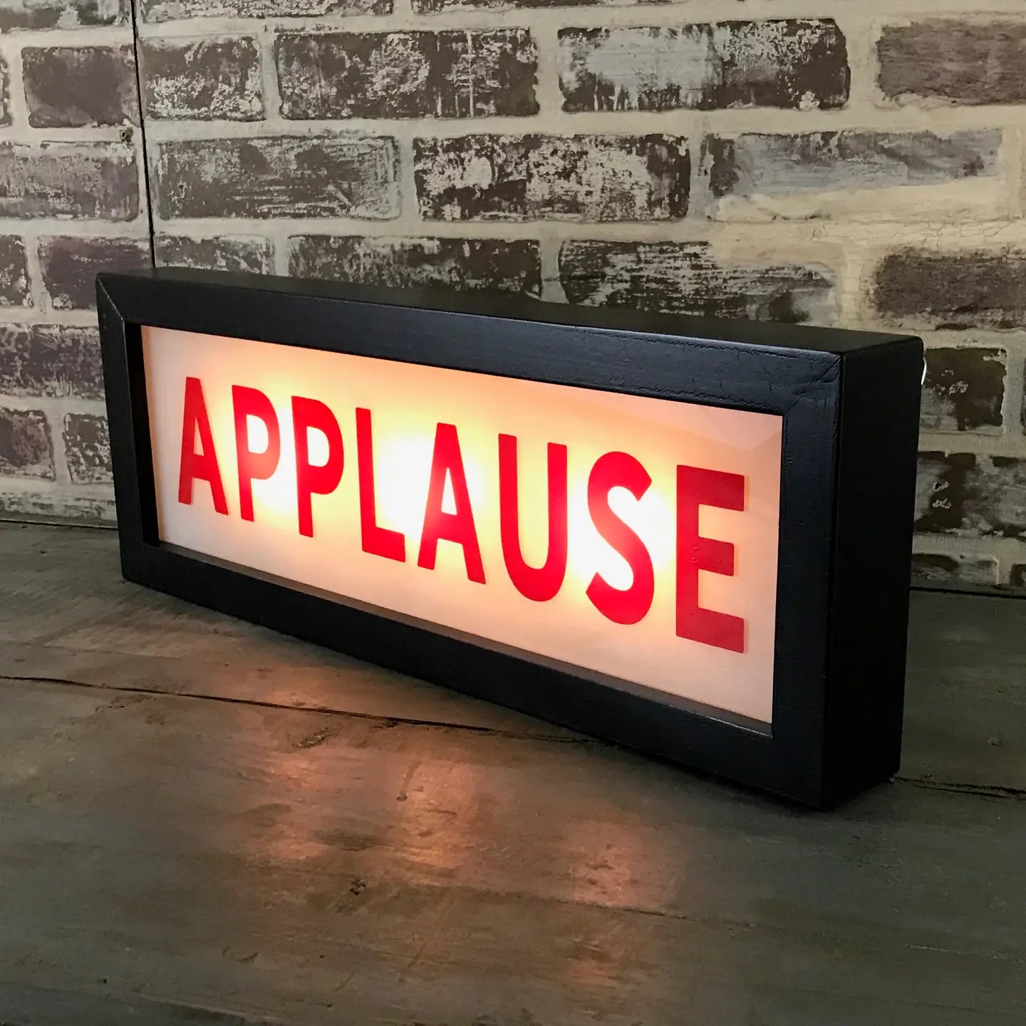 The Faire Lighted Sign is a rectangular lightbox displaying "APPLAUSE" in bold red letters, illuminated against a rustic brick wall background and set on a wooden surface.