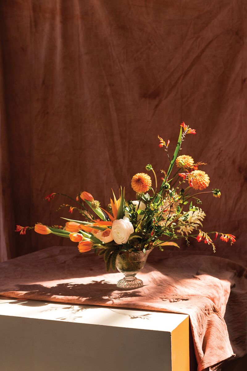 The Camilla Compote by Accent Decor, a glass vase with a 5.5-inch opening, displays orange tulips, yellow and orange flowers, and greenery on a pink-draped table against a soft brown backdrop in warm sunlight.