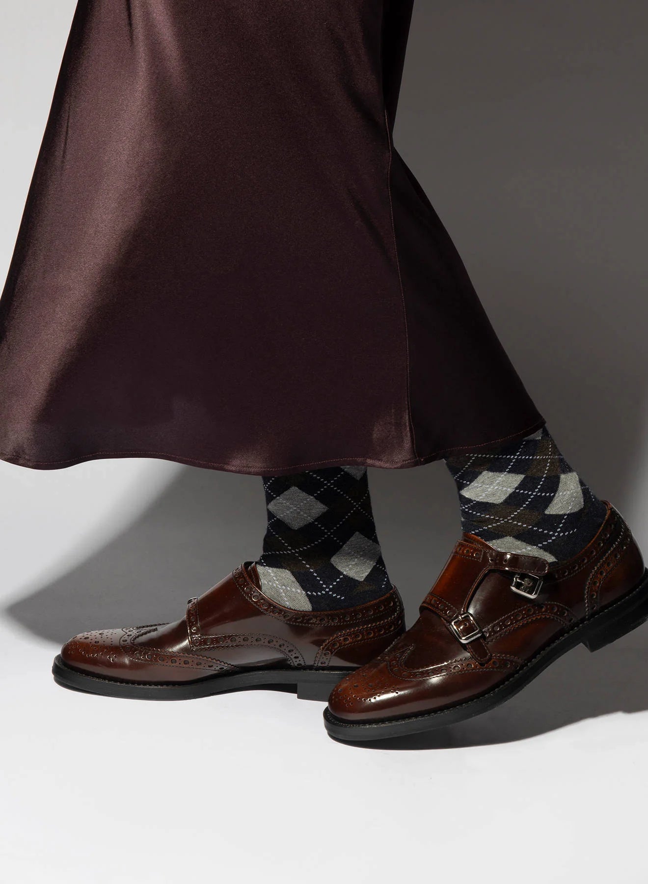 A person in a dark, silky skirt, Maria La Rosa Harlekin Socks, and brown leather monk strap dress shoes stands against a neutral background.