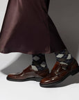 A person in a dark, silky skirt, Maria La Rosa Harlekin Socks, and brown leather monk strap dress shoes stands against a neutral background.