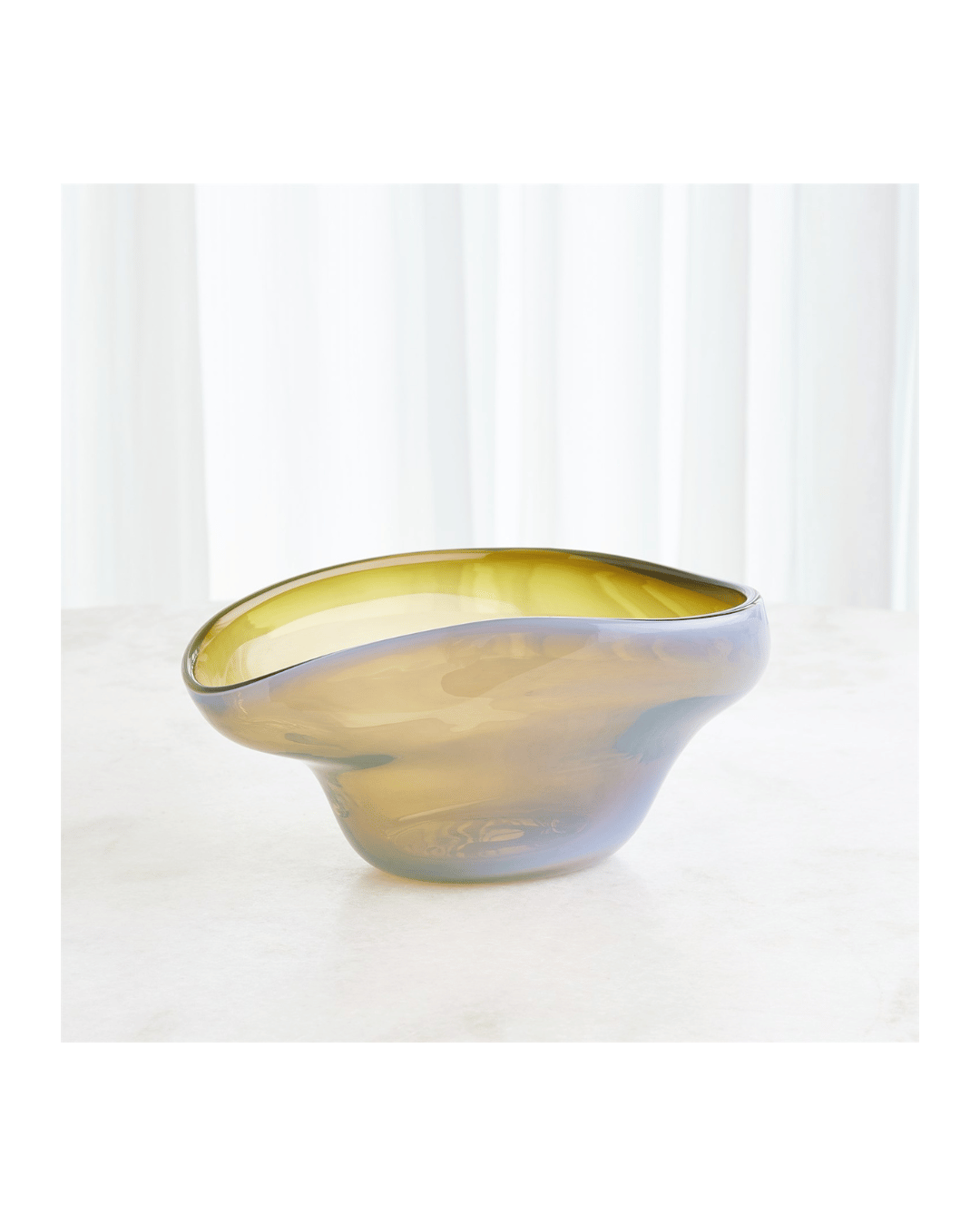 A translucent, irregularly shaped glass bowl with an olive tint sits on a light surface. This exquisite piece of handblown art glass from Global Views complements the soft, white vertical drapes in the background, creating an elegant setting reminiscent of Harmony Bowls.