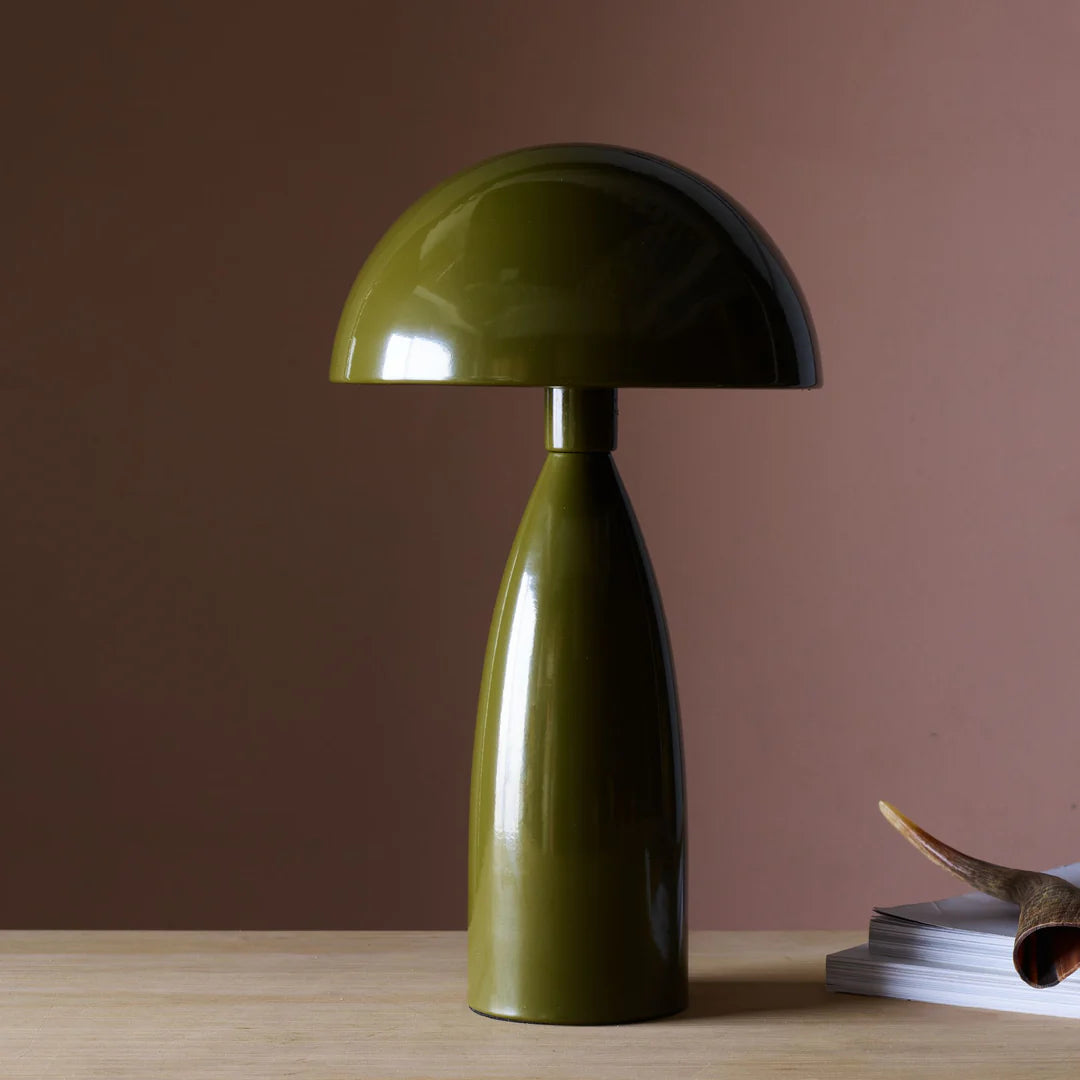 The Leoma Lamp Olive by Abigail Ahern, with its modern dome shade in olive green, sits on a light wooden surface beside a stack of books and an antler against a muted brown wall.