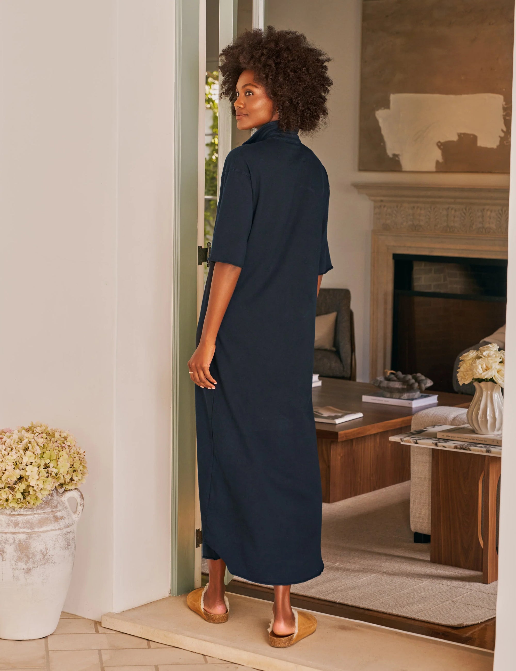 A woman with curly hair stands barefoot in a doorway, wearing the Frank & Eileen Emma Popover Henley Dress in navy. She looks over her shoulder, revealing a cozy living room behind her.
