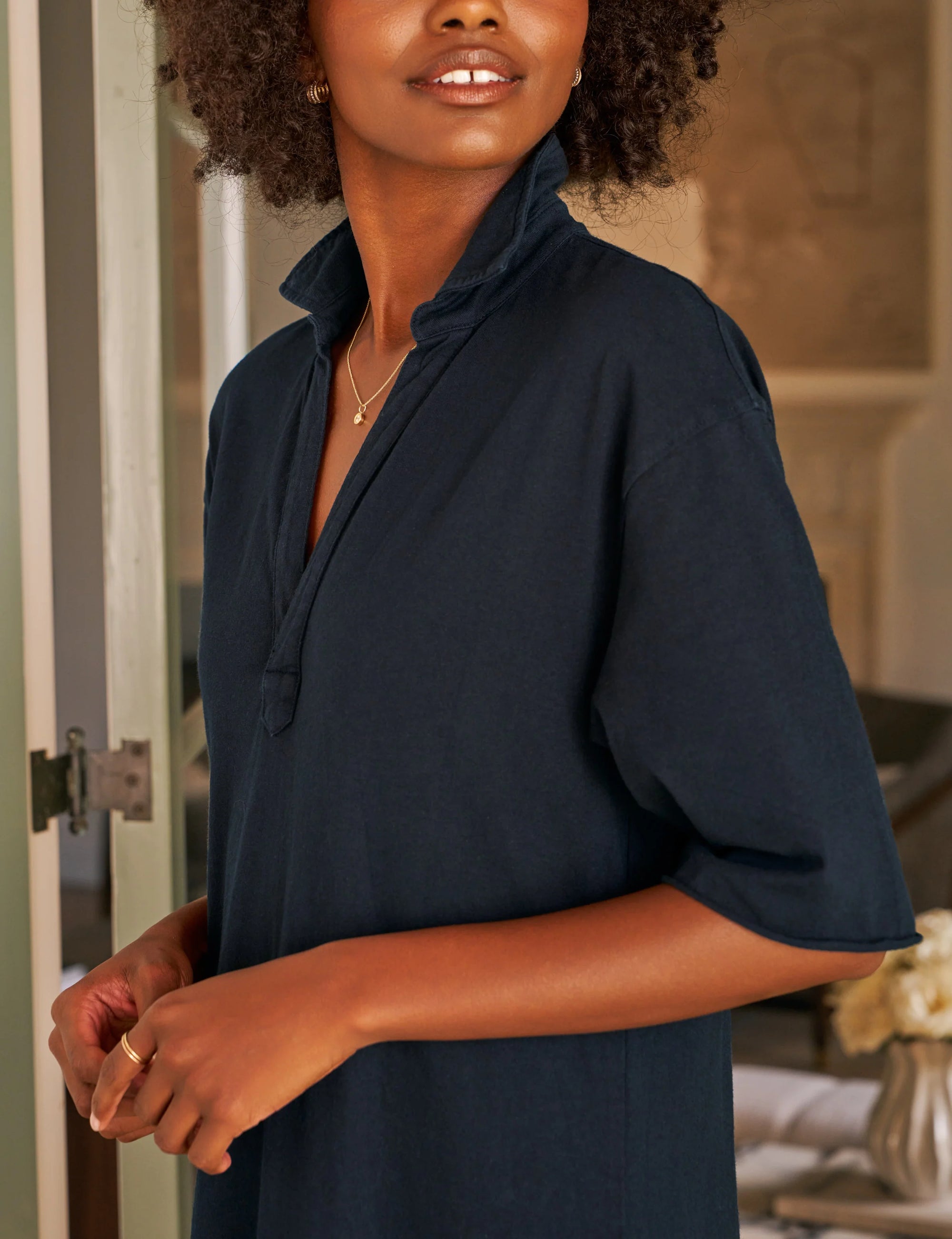 A woman with curly hair wears the Emma Popover Henley Dress by Frank & Eileen and delicate gold jewelry, standing indoors near an open door as soft natural light highlights her face and hands.