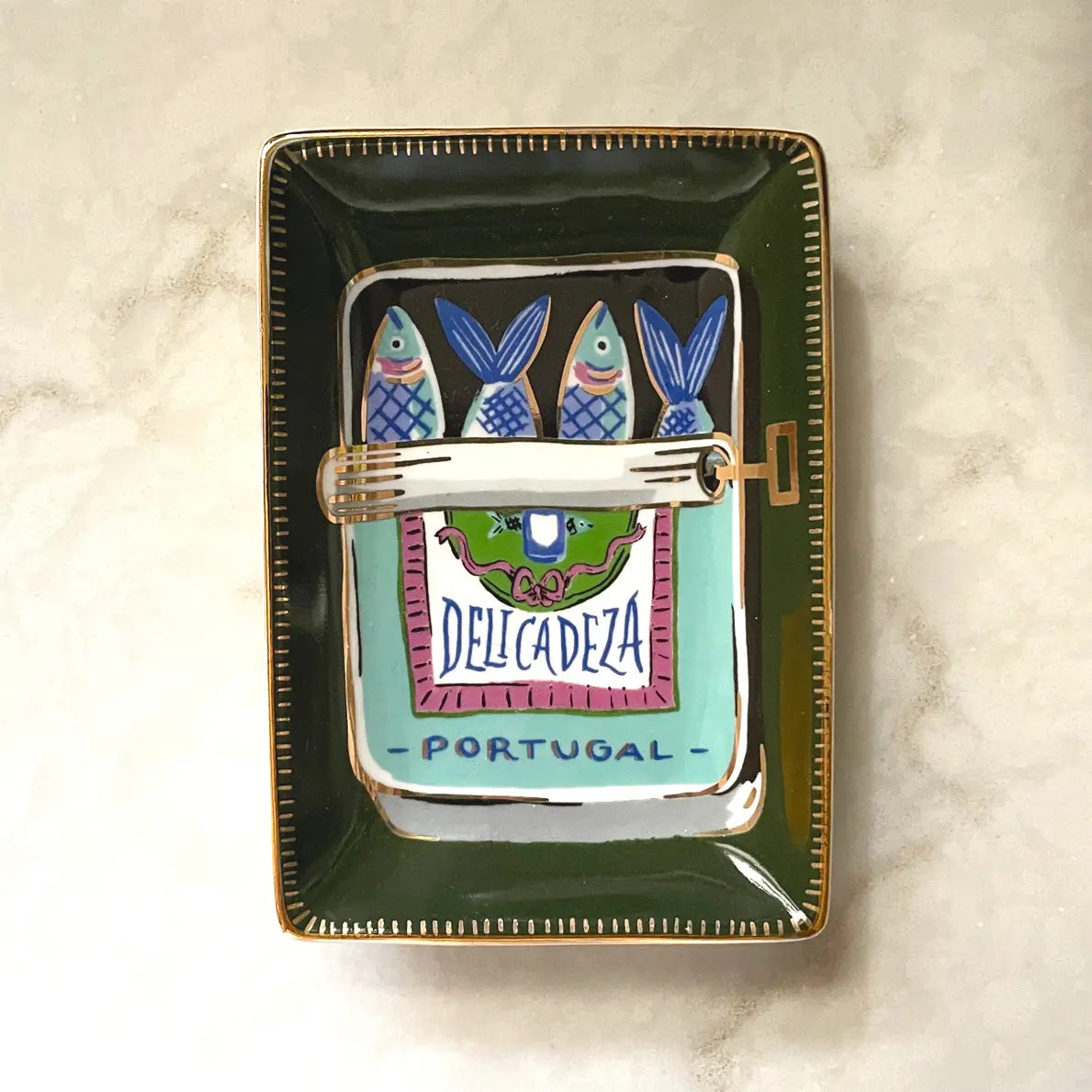 The Faire Trinket Dish is a decorative ceramic piece featuring hand-painted designs with three blue fish and a vibrant label that says "Delicadeza Portugal." It has a green border accentuated with metallic gold details, beautifully displayed on a light marble surface.