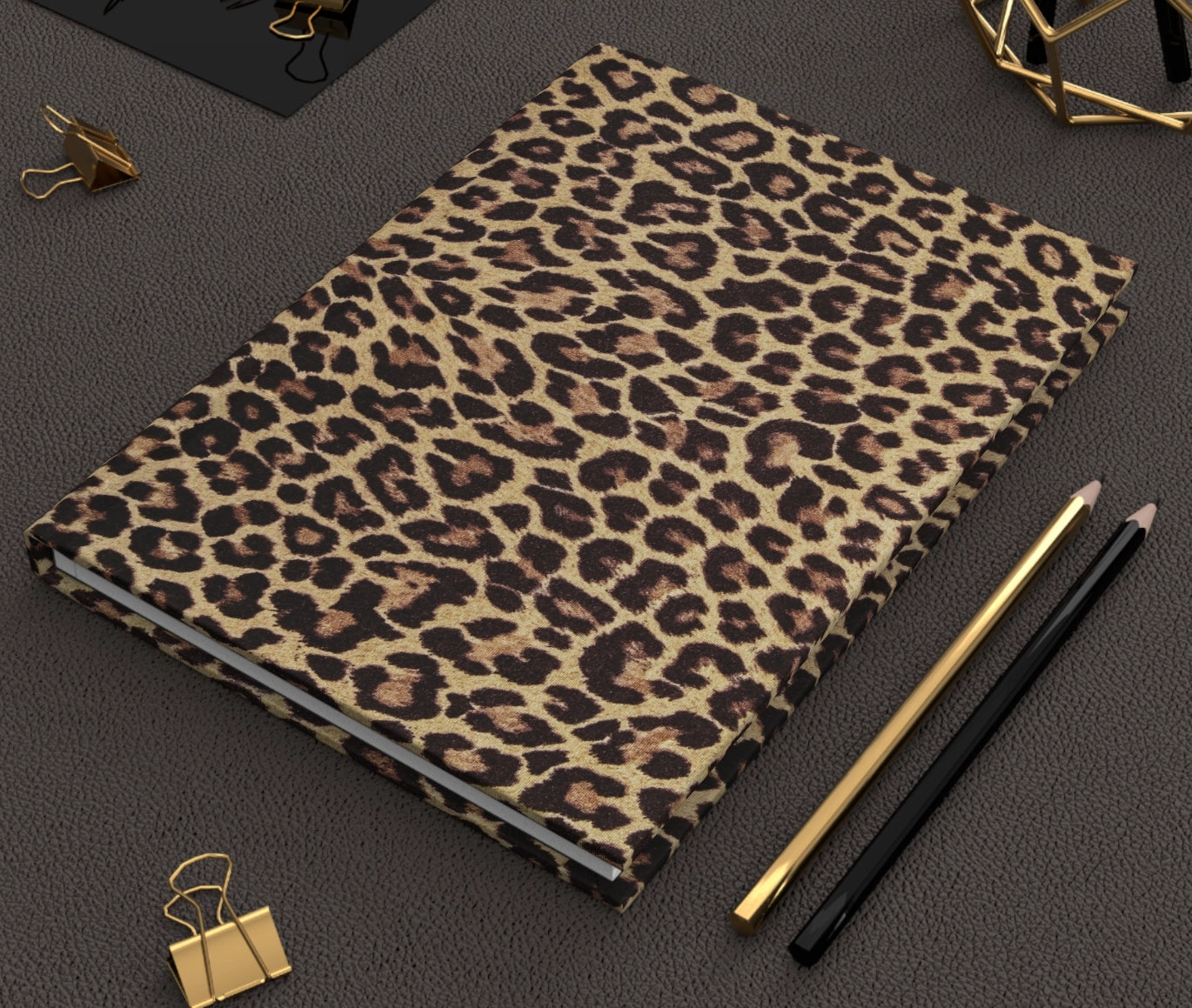 The Faire Leopard Journal with a hardcover design rests on a textured dark surface beside gold and black pencils, gold binder clips, and geometric desk decor.