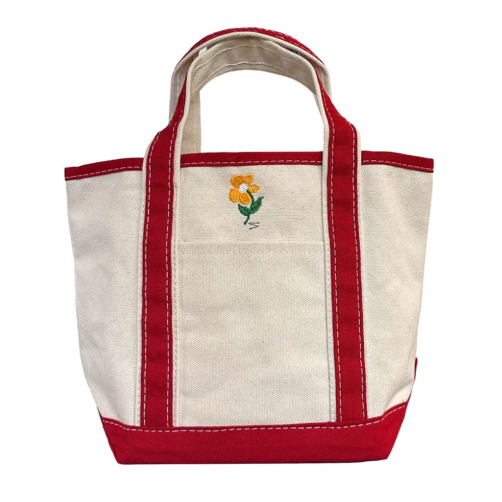 The Faire Deerest Darlin' canvas tote bag is cream-colored with red handles and trim, and has an embroidered yellow flower with green leaves on the front pocket.