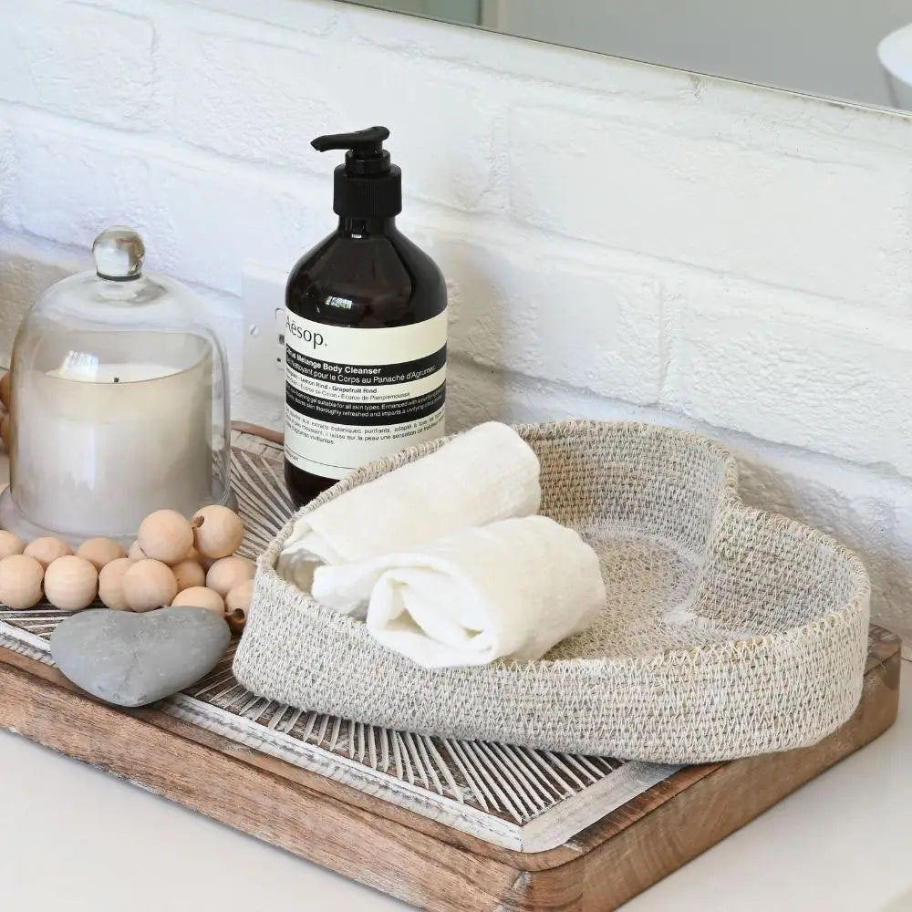 On a bathroom countertop against a white brick wall, two rolled white towels rest in the Indaba Heart Seagrass Basket alongside a tray with a candle, wooden beads, a heart-shaped stone, and a soap dispenser.