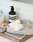 On a bathroom countertop against a white brick wall, two rolled white towels rest in the Indaba Heart Seagrass Basket alongside a tray with a candle, wooden beads, a heart-shaped stone, and a soap dispenser.