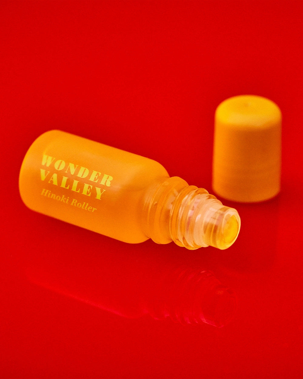 A small yellow bottle labeled "Faire Hinoki Roll On" rests open on a vibrant red surface, its matching cap nearby—ready to release a calming, plant-based hinoki cedar fragrance.