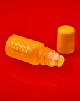 A small yellow bottle labeled "Faire Hinoki Roll On" rests open on a vibrant red surface, its matching cap nearby—ready to release a calming, plant-based hinoki cedar fragrance.