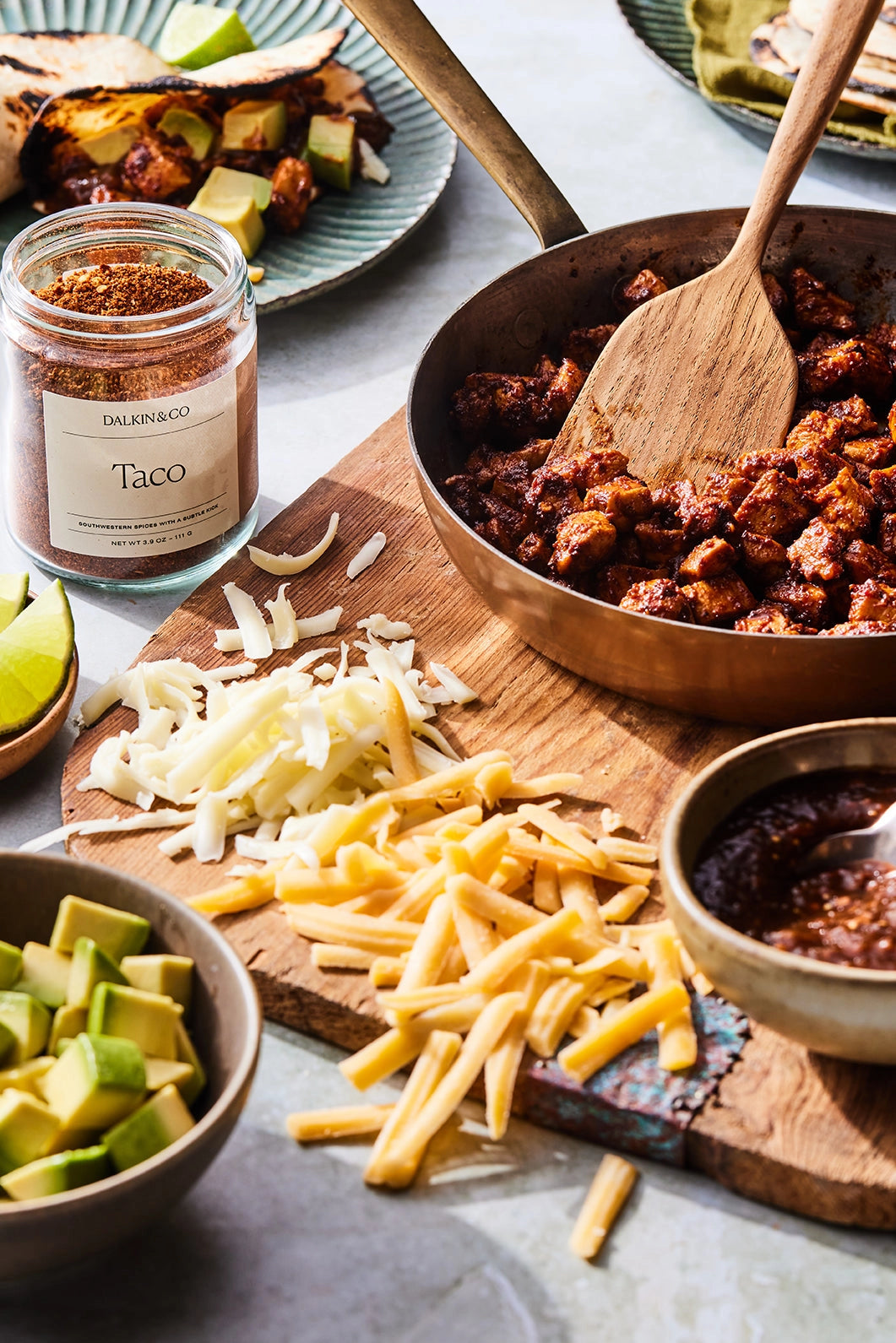 A wooden board with shredded cheese, diced avocado, seasoned taco meat, salsa, lime wedges, and a jar of Faire Taco Seasoning adds bold Southwestern flavor to this fresh tortilla wrap spread.