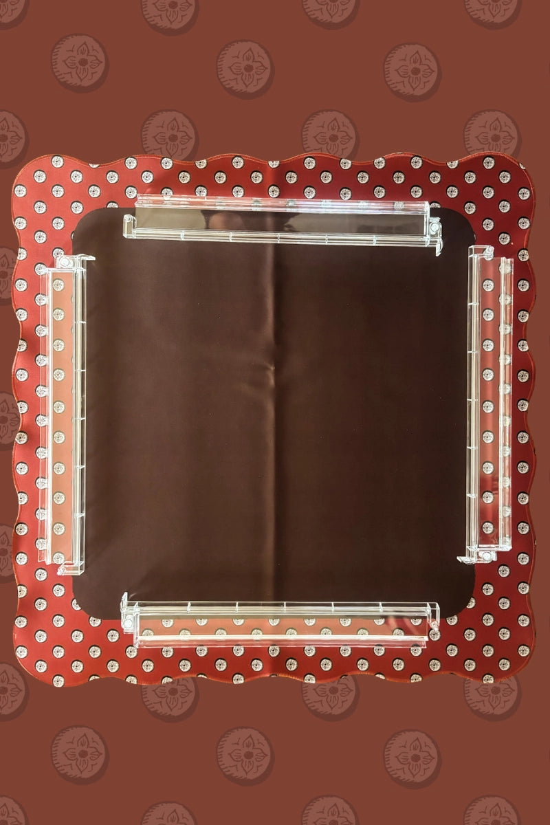 The Wild West Mahjong Mat ONLY by Faire is a square, dark brown mat with scalloped red edges and white circle patterns, topped with four transparent acrylic rods—one per side—and set against a matching patterned backdrop.