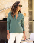 A woman with curly hair and sunglasses stands outside in a stylish Frank & Eileen Patrick Popover Henley top paired with white pants. She exudes preppy flair while posing by a stone wall, with part of a fireplace visible in the background.