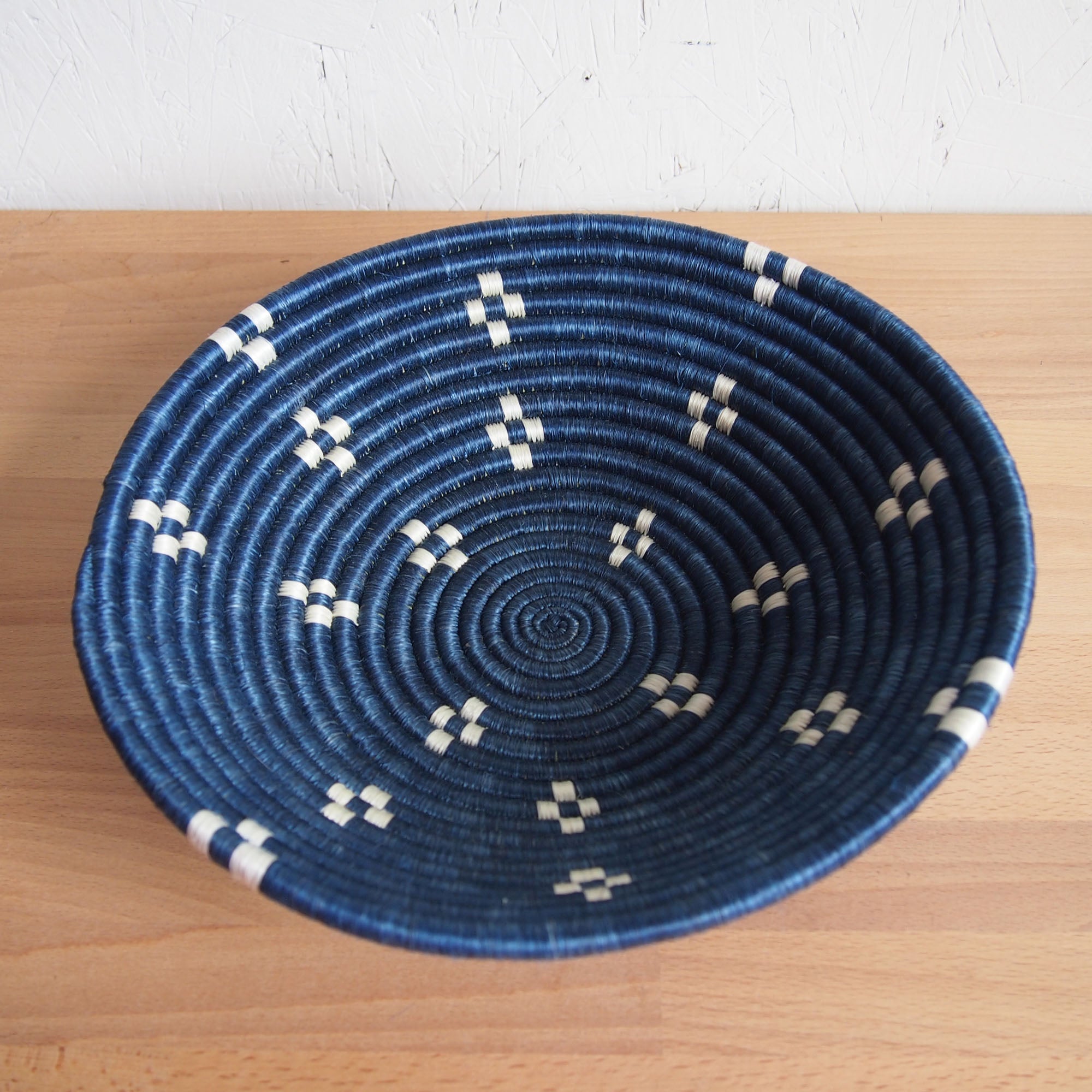 The Kabaya Large Bowl by Amsha - Faire is a round blue sisal woven basket with a coiled design and small white geometric patterns on both sides, shown on a light wooden surface.