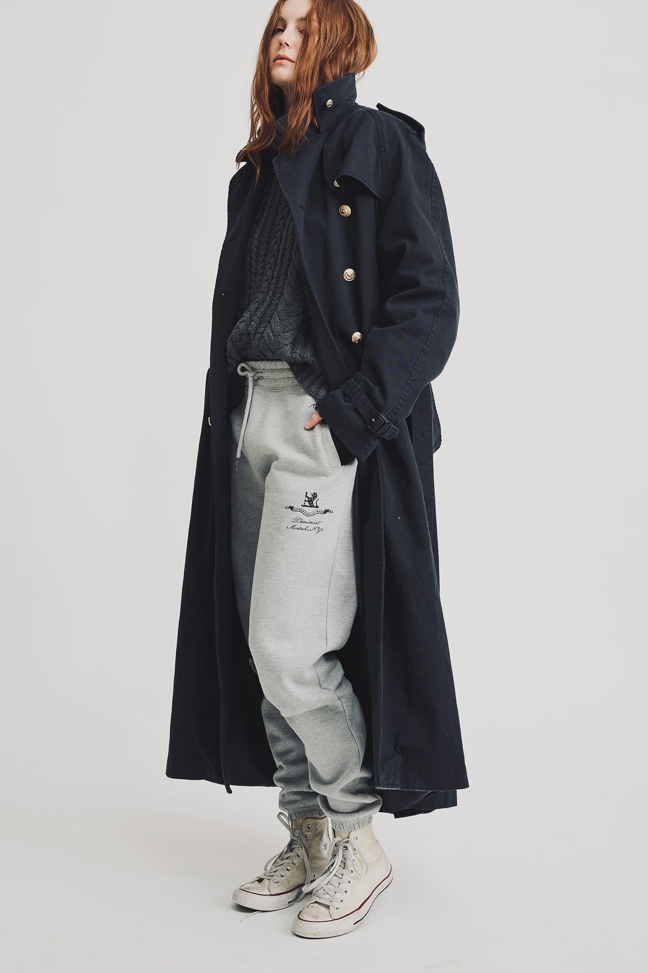 A person with long red hair wears the Denimist OVERSIZED TRENCH COAT, a gray cable-knit sweater, gray sweatpants, and white high-top sneakers, standing against a plain white background.
