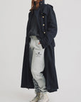 A person with long red hair wears the Denimist OVERSIZED TRENCH COAT, a gray cable-knit sweater, gray sweatpants, and white high-top sneakers, standing against a plain white background.
