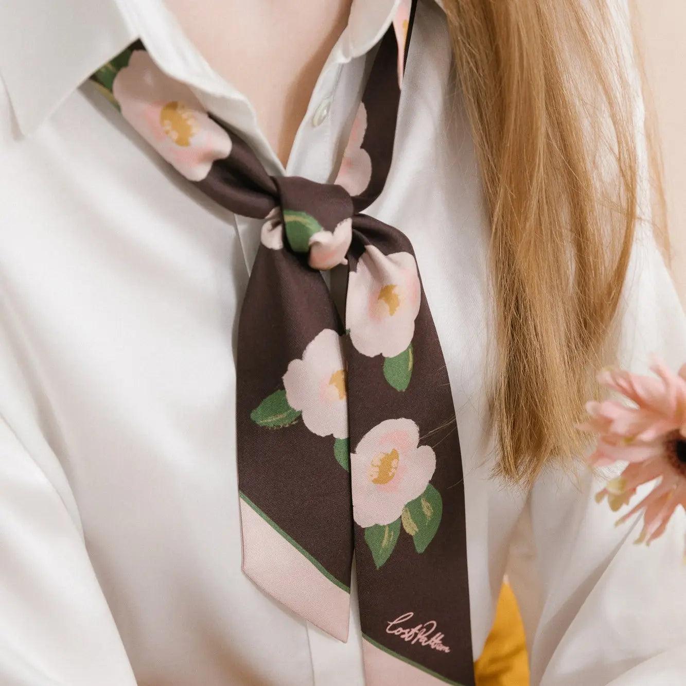 A person wearing a white blouse accessorized with the Camellia Silk Twilly Neck Bow Dark Brown by Faire, styled into a graceful bow at the neck. The person's long hair drapes elegantly over their shoulder, and they hold a pink flower near the bottom right of the image.