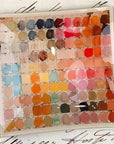 The Faire Watercolor Artist's Palette Glass Tray features a grid of vibrant watercolor swatches—blues, pinks, reds, oranges, yellows, and browns—overlaid on paper with elegant cursive script for an inspiring artful accent.