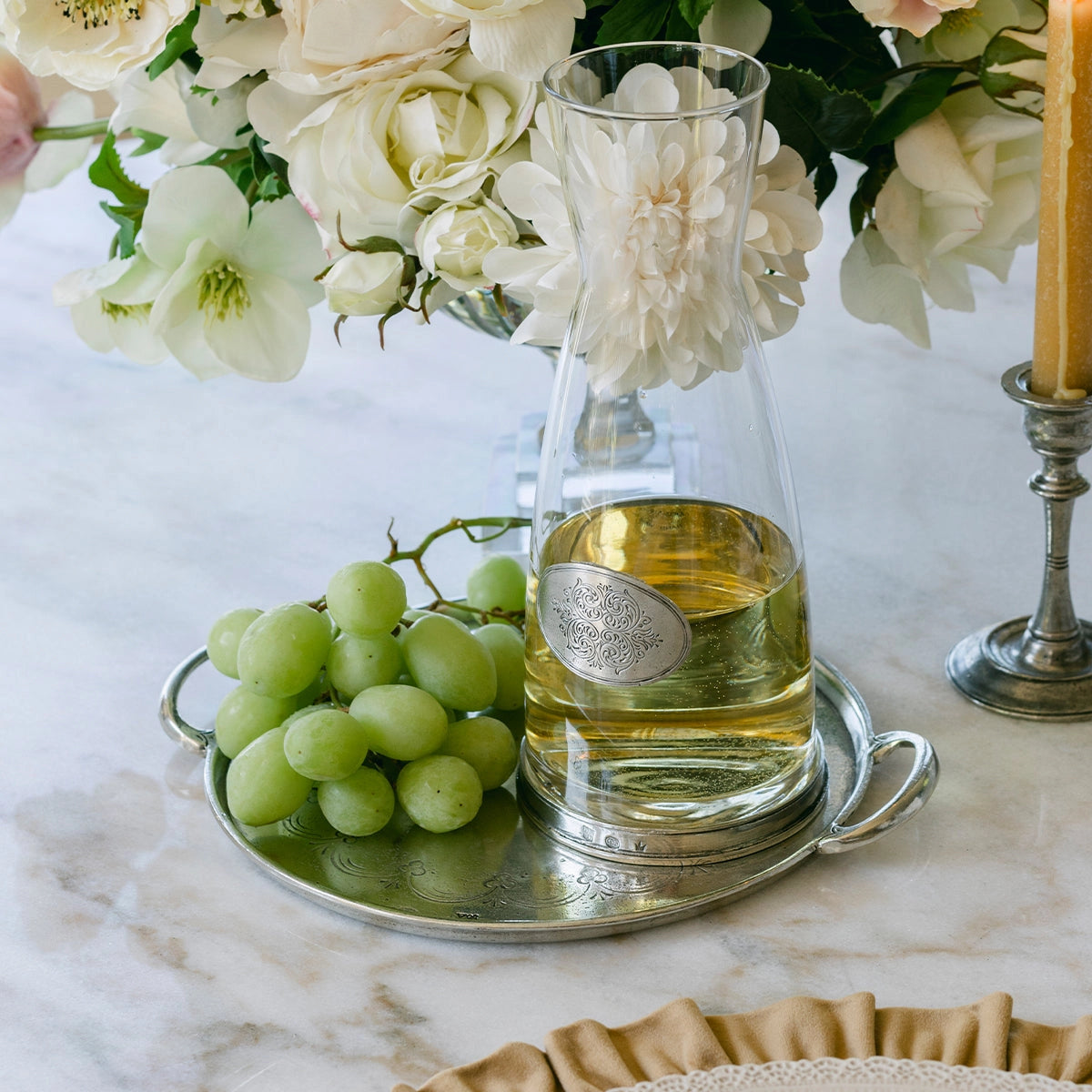 A glass carafe with white wine rests on the Faire Vintage Pewter Round Tray With Handles beside green grapes, with white flowers and a candle in the background atop a marble table.