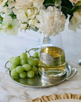 A glass carafe with white wine rests on the Faire Vintage Pewter Round Tray With Handles beside green grapes, with white flowers and a candle in the background atop a marble table.