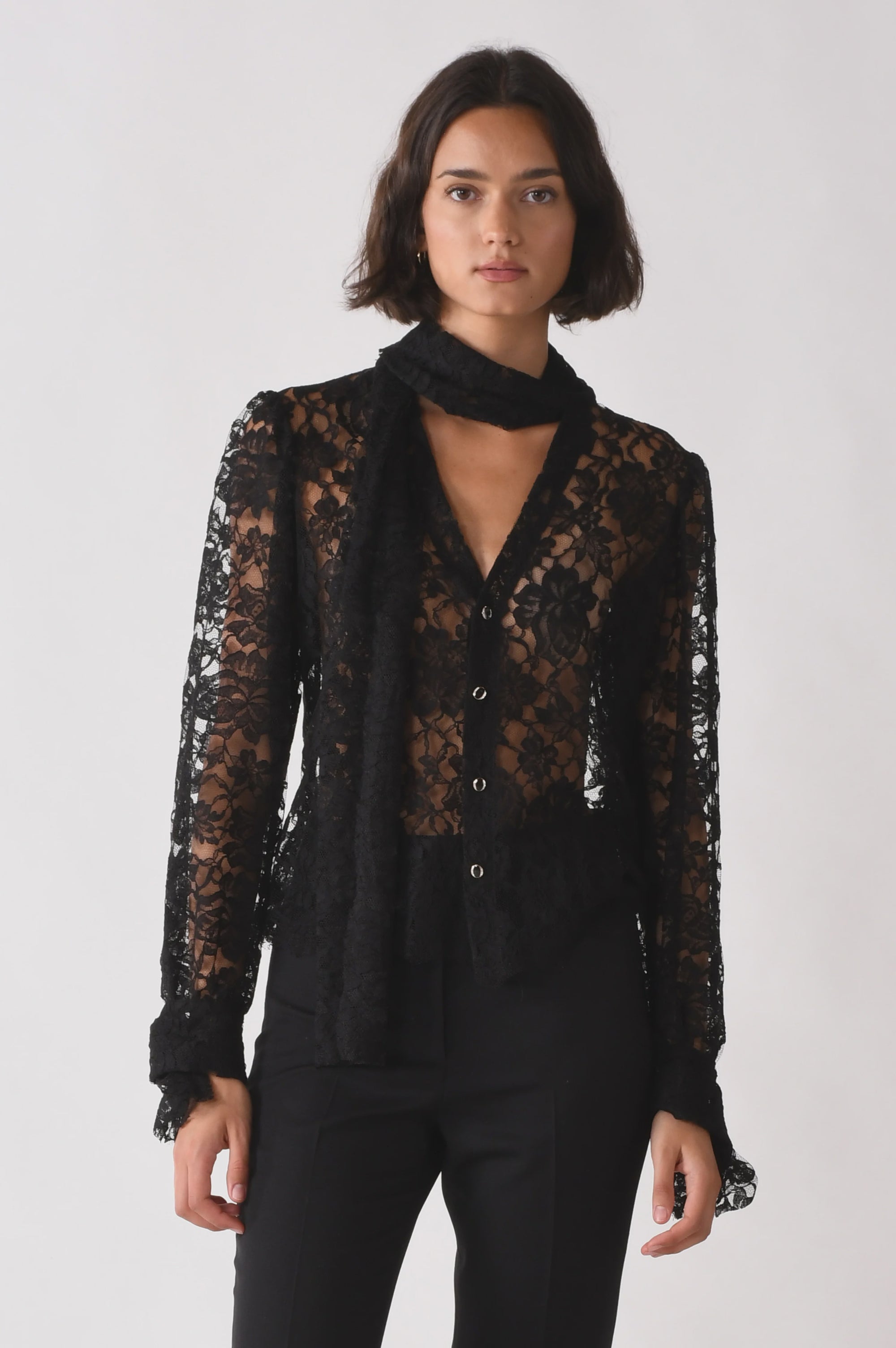 A woman wears The Bow Tie Shirt by Cissa—a sheer black lace blouse with long sleeves and a scalloped hem—paired with black pants and a matching lace scarf, standing against a plain white background.