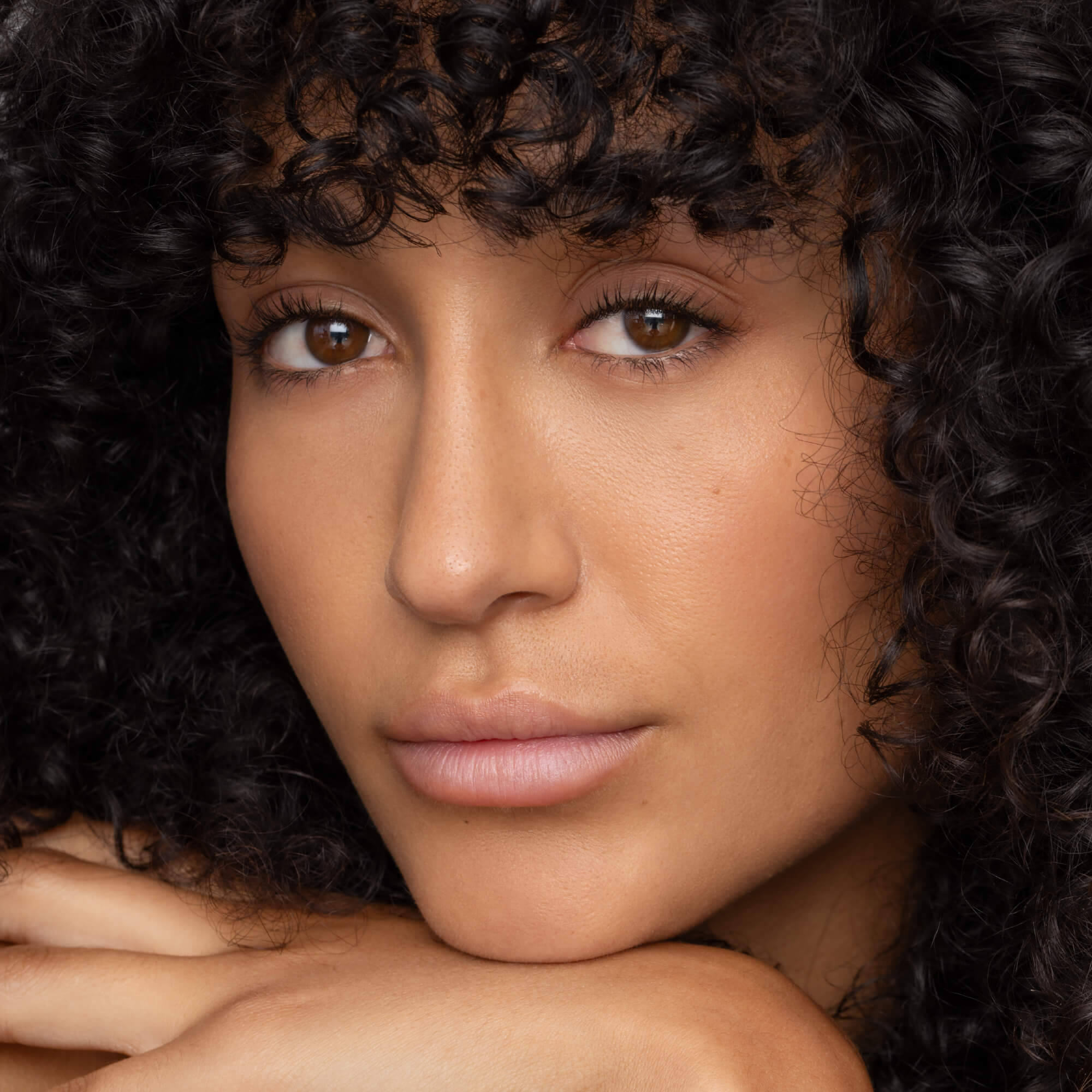 A woman with curly dark hair and brown eyes rests her chin on her hands, gazing calmly at the camera. Her smooth, natural skin is complemented by minimal makeup and a hint of Faire’s Poppy & Pout Lip Tint in Farrah.