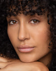 A woman with curly dark hair and brown eyes rests her chin on her hands, gazing calmly at the camera. Her smooth, natural skin is complemented by minimal makeup and a hint of Faire’s Poppy & Pout Lip Tint in Farrah.