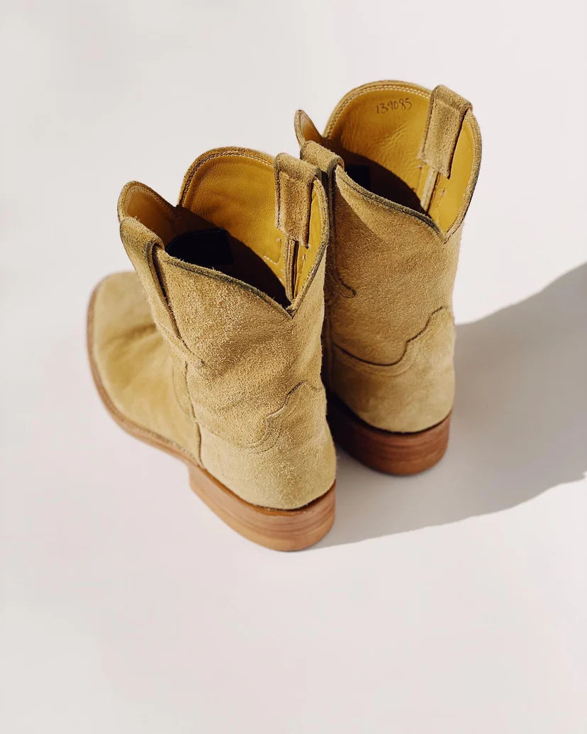 A pair of "The Ranch Boot" by Hey Gang, crafted from tan calf skin rough out suede with wooden heels, is viewed from the back on a light surface with shadows cast to the side.