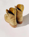 A pair of "The Ranch Boot" by Hey Gang, crafted from tan calf skin rough out suede with wooden heels, is viewed from the back on a light surface with shadows cast to the side.