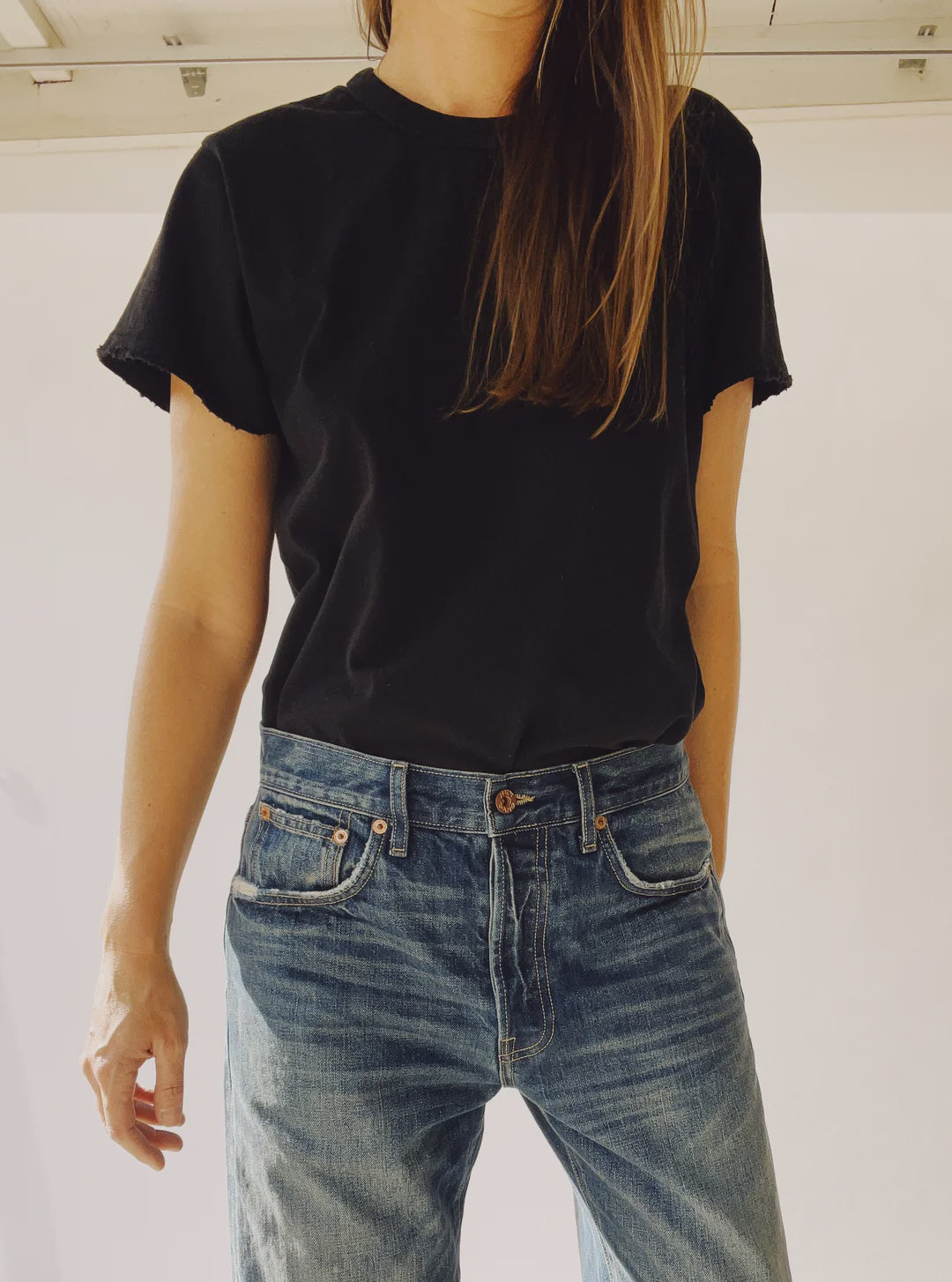 A person wearing "The Crew,” a vintage-fit black cotton T-shirt by Hey Gang, along with blue jeans, stands in front of a white background, their long hair partially covering their face. Made in Los Angeles, the outfit exudes casual style with hands relaxed at their sides.