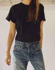 A person wearing "The Crew,” a vintage-fit black cotton T-shirt by Hey Gang, along with blue jeans, stands in front of a white background, their long hair partially covering their face. Made in Los Angeles, the outfit exudes casual style with hands relaxed at their sides.