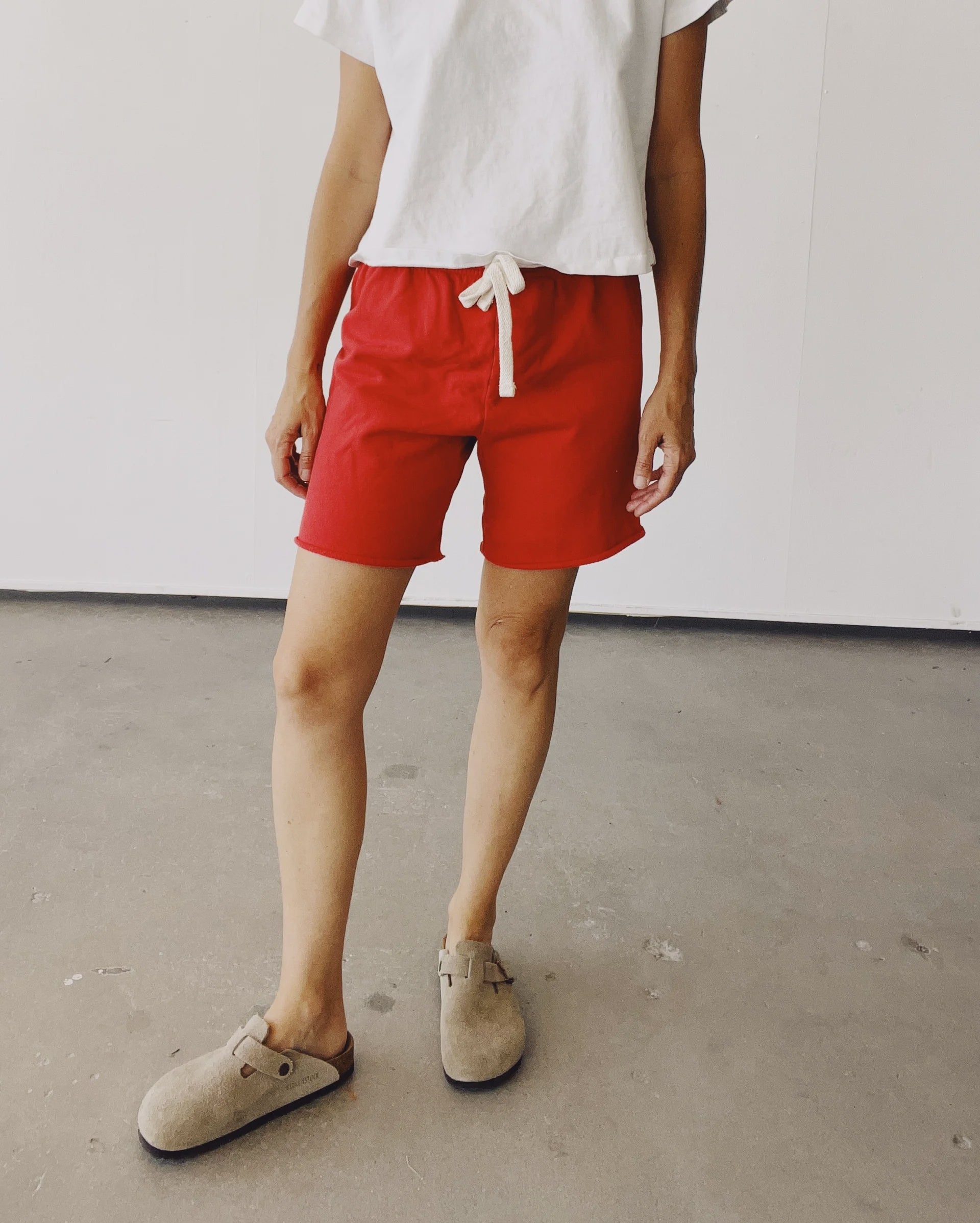 A person stands indoors wearing Hey Gang’s The Sweat Shorts in vintage red with a white drawstring, paired with a white t-shirt and light tan slip-on shoes, against a minimalistic background.