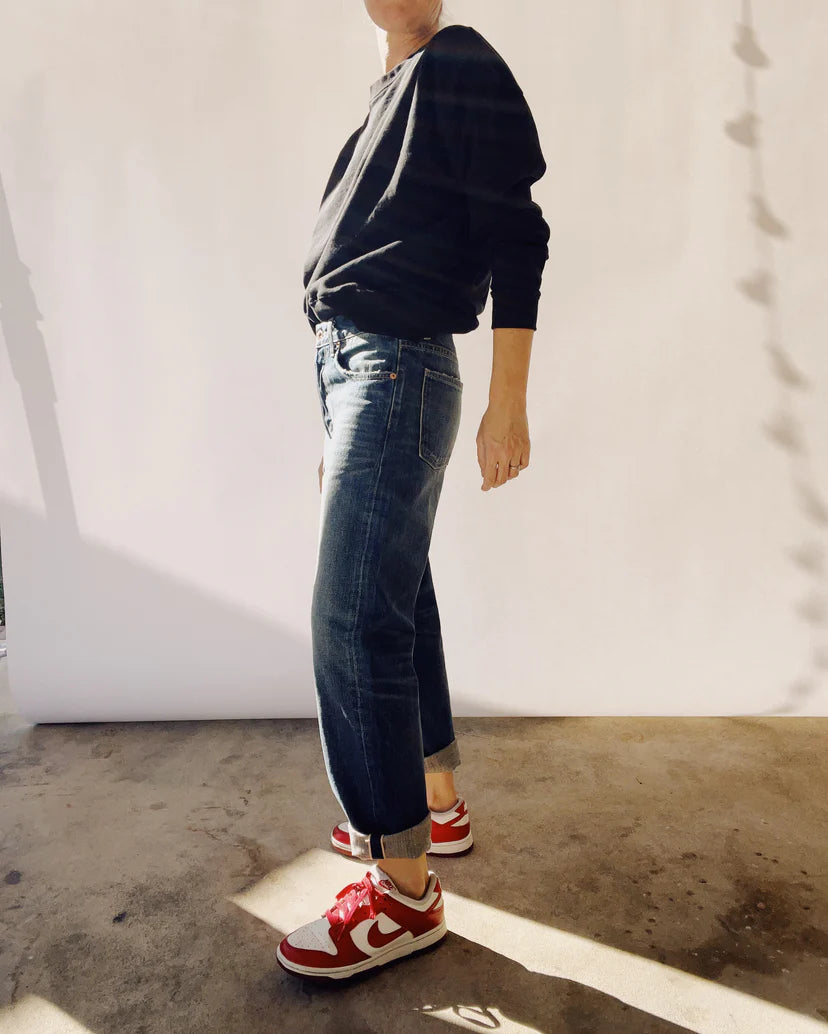 A person wearing a black long-sleeve shirt, The Loose Crop Selvedge jeans by Hey Gang rolled up at the cuffs, and red and white sneakers stands in sunlight on a concrete floor, casting shadows on a white background.