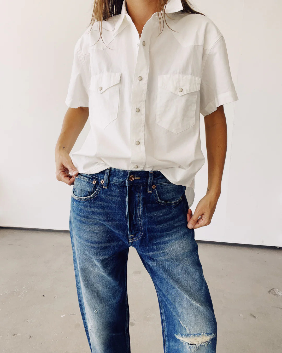 A person wearing &quot;The Western Shirt&quot; by Hey Gang, featuring chest pockets, along with blue distressed jeans, stands against a plain background. Their face is not visible.