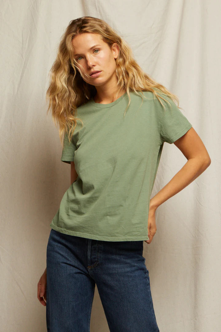 A woman with long wavy blonde hair wears the Perfectwhitetee Harley SS Boxy Crew in light green and mid-rise denim. She stands before a plain beige fabric backdrop, one hand in her pocket, looking at the camera.