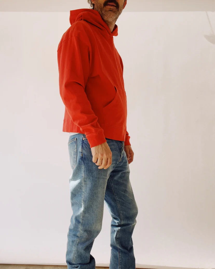 A person stands sideways wearing The Hoodie from Hey Gang, which is designed in a unisex fit and features a vibrant red color. Paired with blue jeans against a plain, light background, this attire crafted from 100% cotton showcases a casual and relaxed style as the individual casually rests their hands in their pockets.