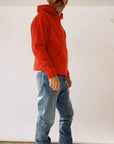 A person stands sideways wearing The Hoodie from Hey Gang, which is designed in a unisex fit and features a vibrant red color. Paired with blue jeans against a plain, light background, this attire crafted from 100% cotton showcases a casual and relaxed style as the individual casually rests their hands in their pockets.