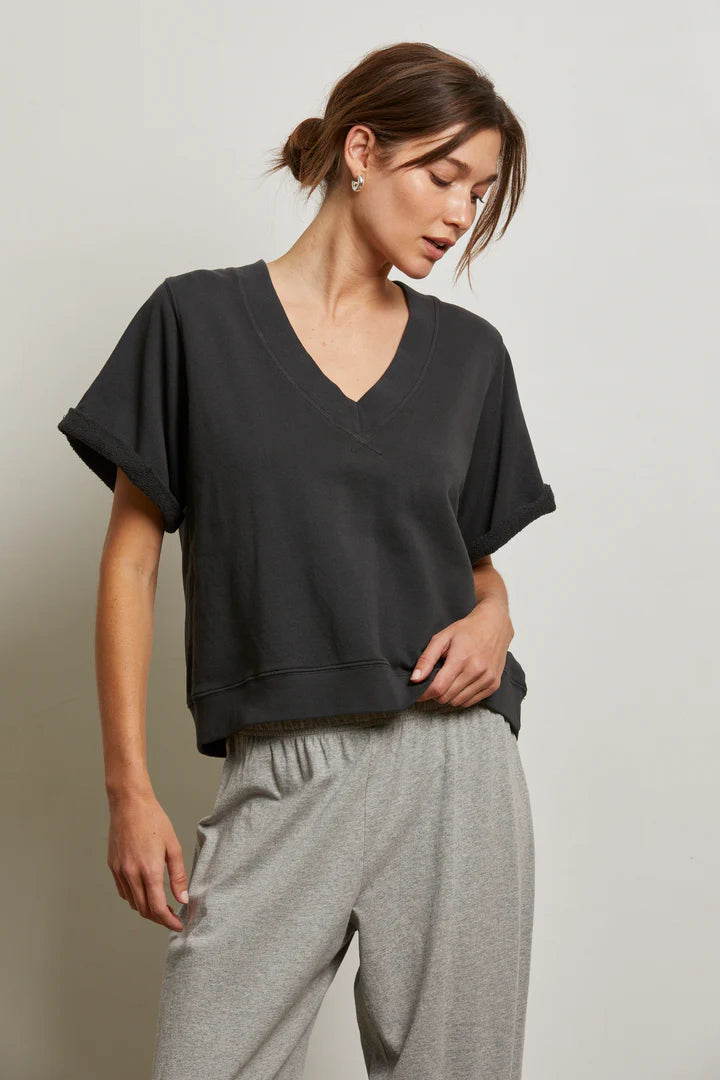 A woman with brown hair in a low bun wears the Perfectwhitetee French Terry SS Crew and light gray drawstring pants, standing against a plain light wall, looking slightly down and to the side.