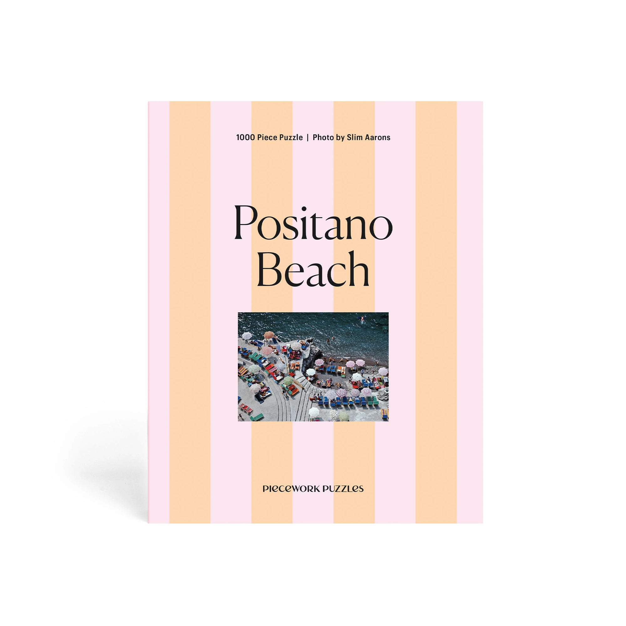 The Faire Pieceworks Puzzle 1000 features pink and peach stripes, "Positano Beach" text, and a small photo of colorful umbrellas on a packed Positano beach—evoking Slim Aarons’ iconic, midcentury seaside style.