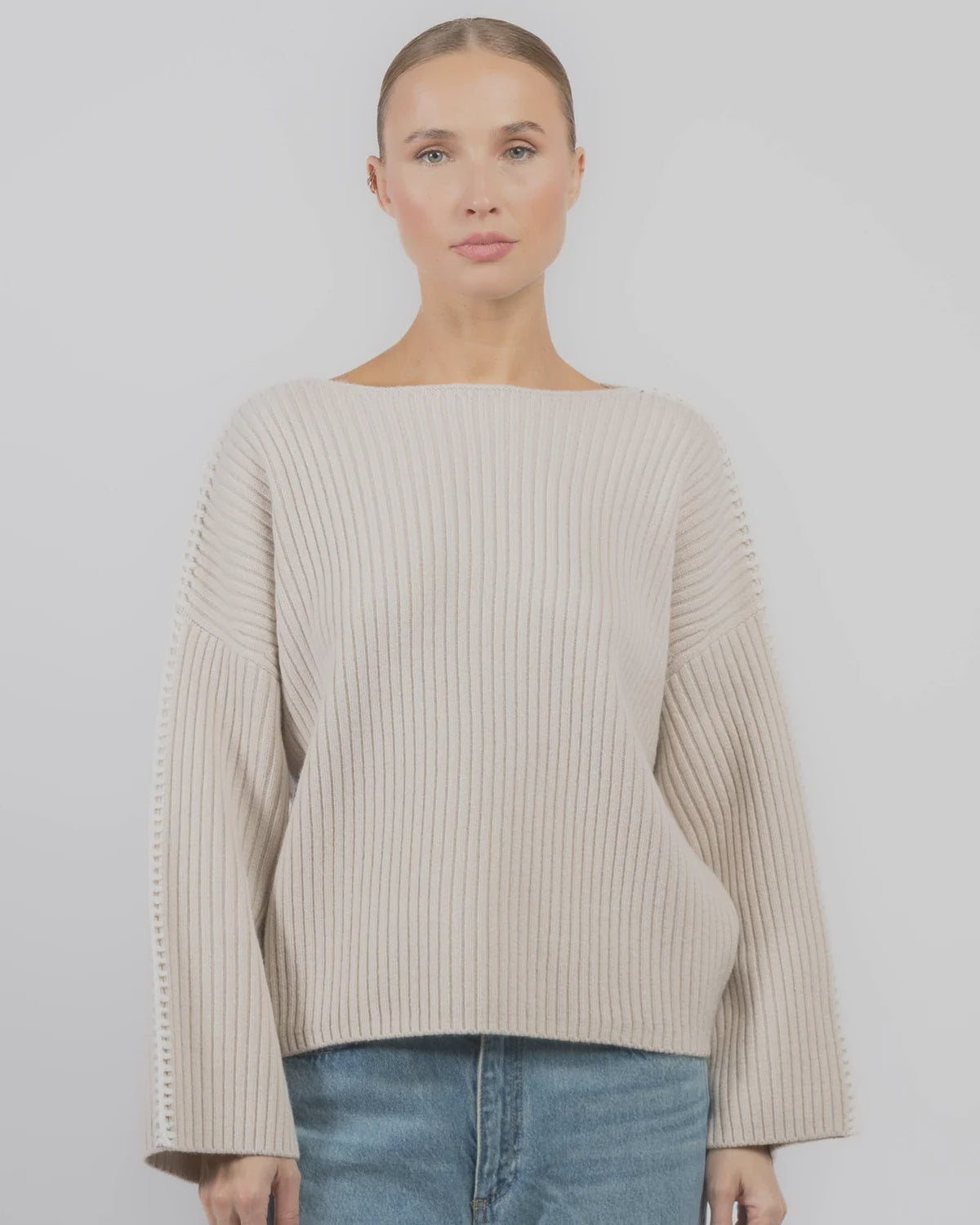 A woman with light hair tied back wears the Rag & Bone ingrid boat neck sweater—loose with chunky ribbing and wide sleeves—paired with blue jeans, standing against a plain light background.
